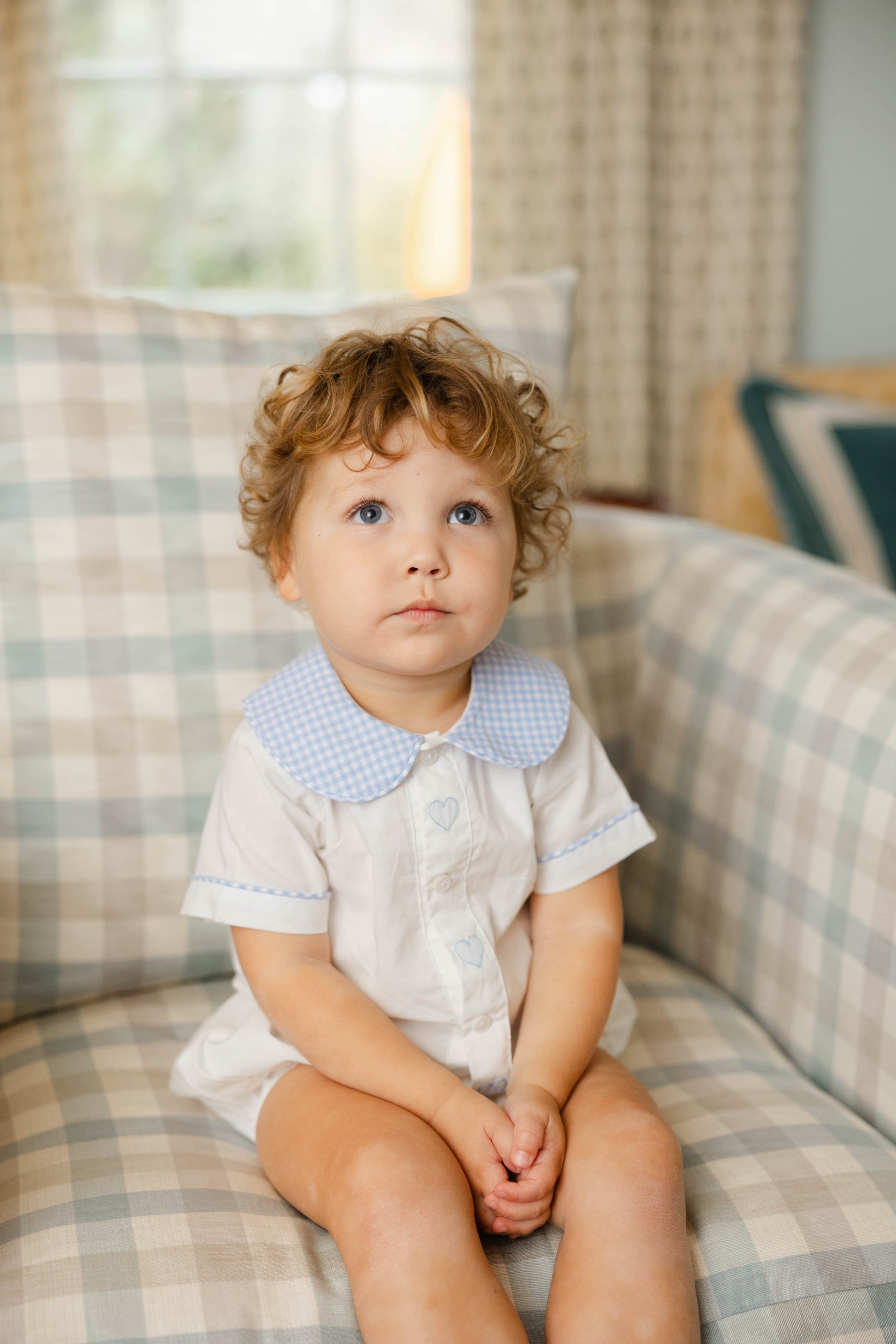 Child sitting on a plaid couch wearing a white outfit with a blue collar.
