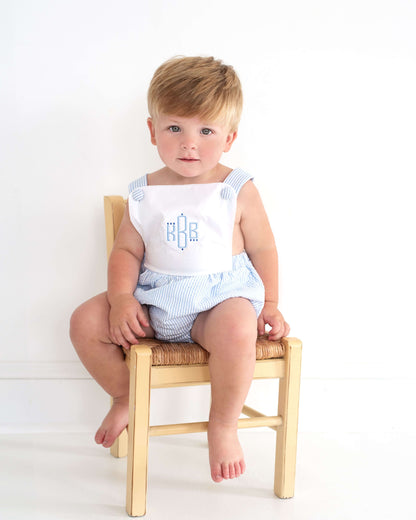 Child sitting on a small wooden chair wearing a white outfit with blue patterns.