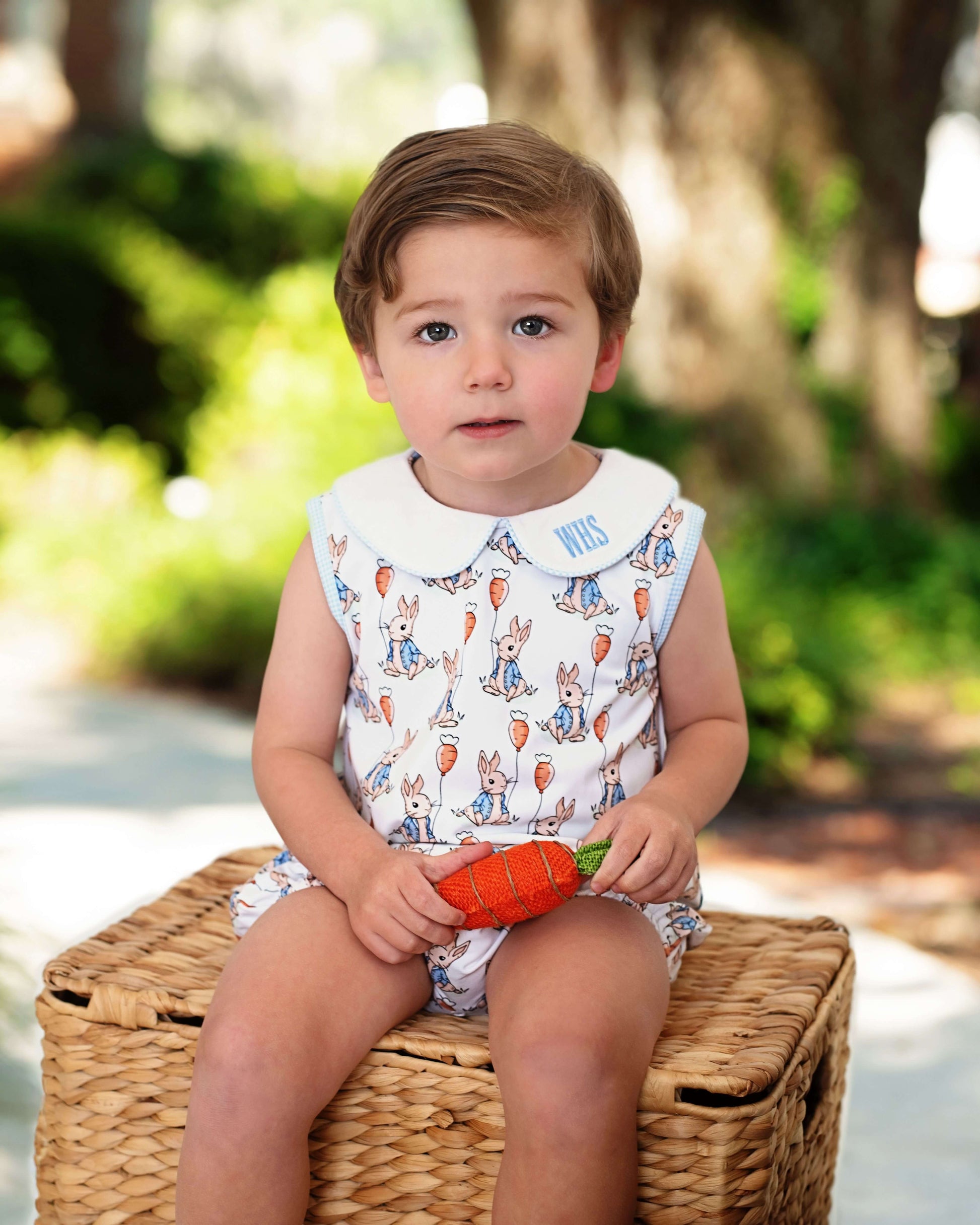 Child sitting on a woven box outdoors holding a carrot toy, wearing a patterned outfit.