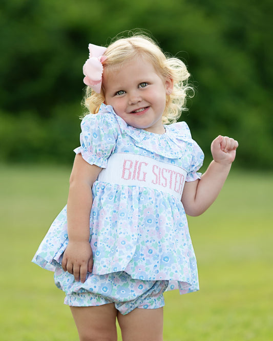 Child wearing a 'BIG SISTER' outfit outdoors with a blurred green background