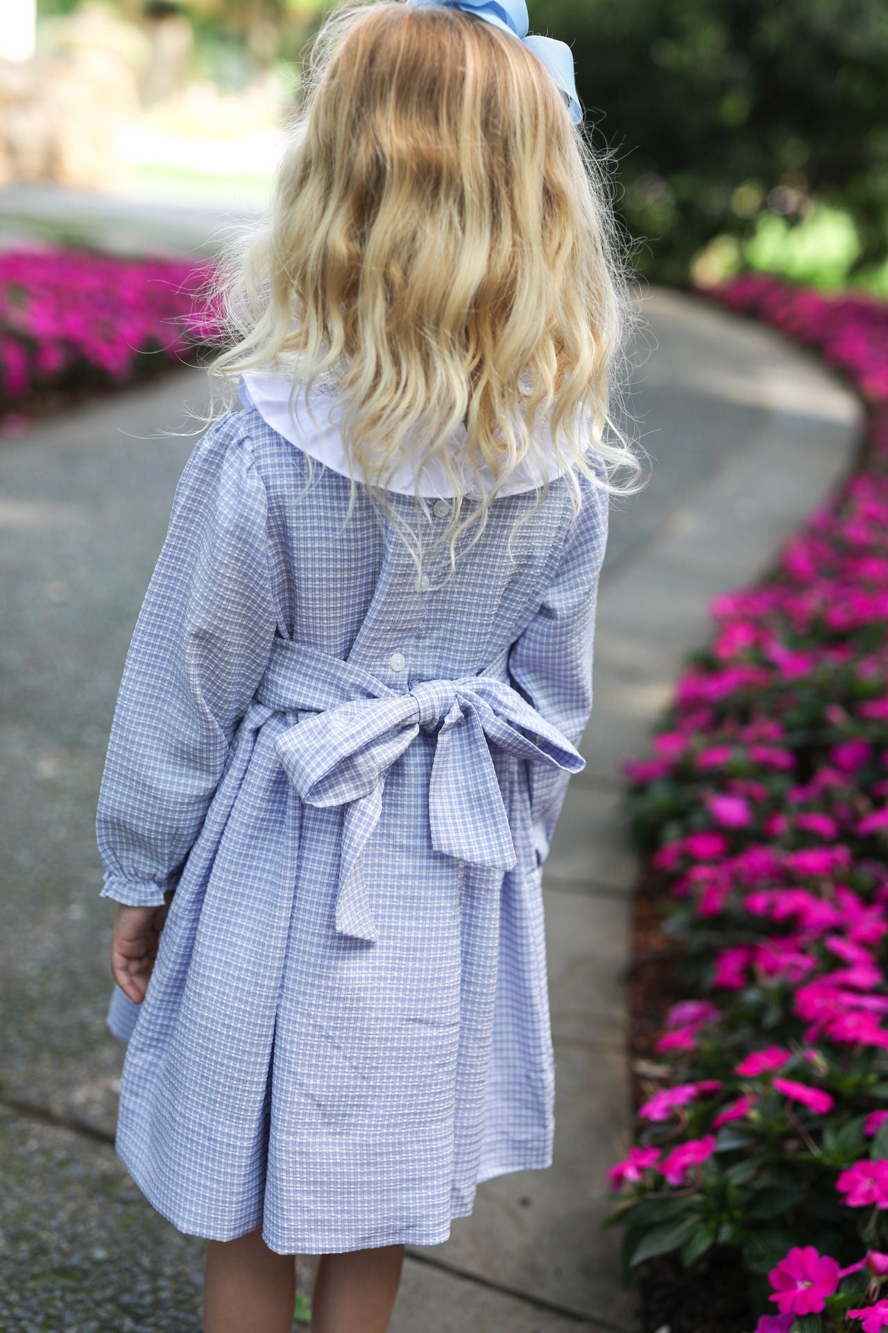 Child wearing a blue checkered dress with a white collar, standing on a path with pink flowers.
