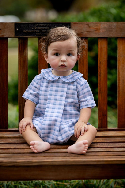 Child wearing a blue checkered outfit sitting on a wooden bench outdoors.