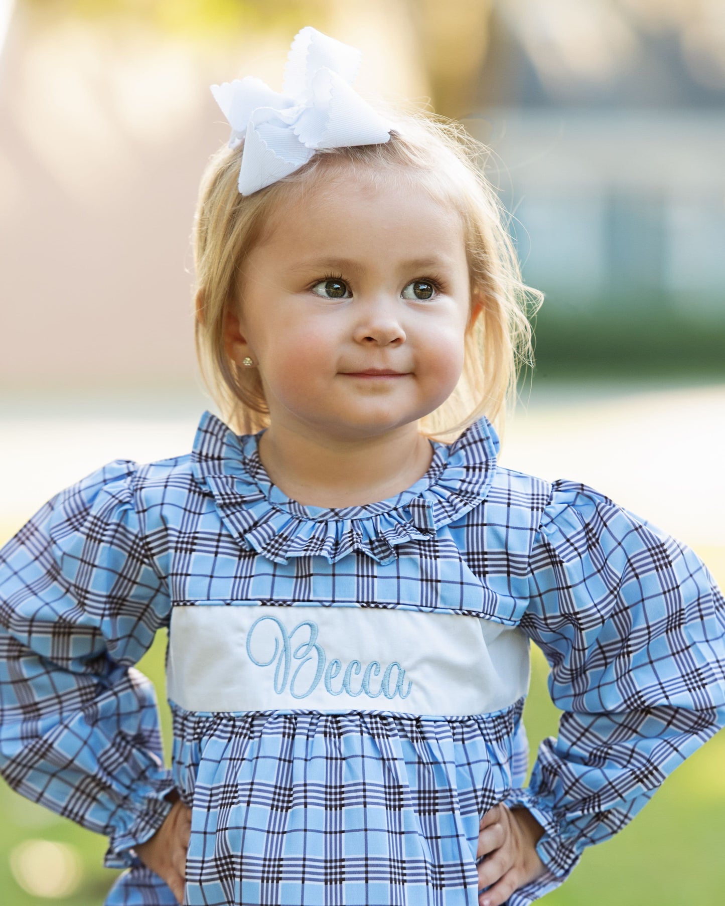 Child wearing a blue plaid dress with 'Becca' embroidered on it, standing outdoors.