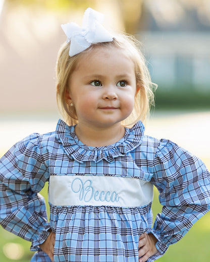 Child wearing a blue plaid dress with 'Becca' embroidered on it, standing outdoors.