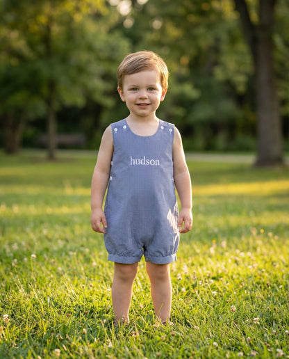 Child wearing a blue romper with 'hudson' printed on it, standing in a grassy field.
