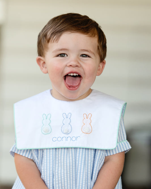Child wearing a blue seersucker bubble with embroidered bunnies and the name 'Connor' on a neutral background