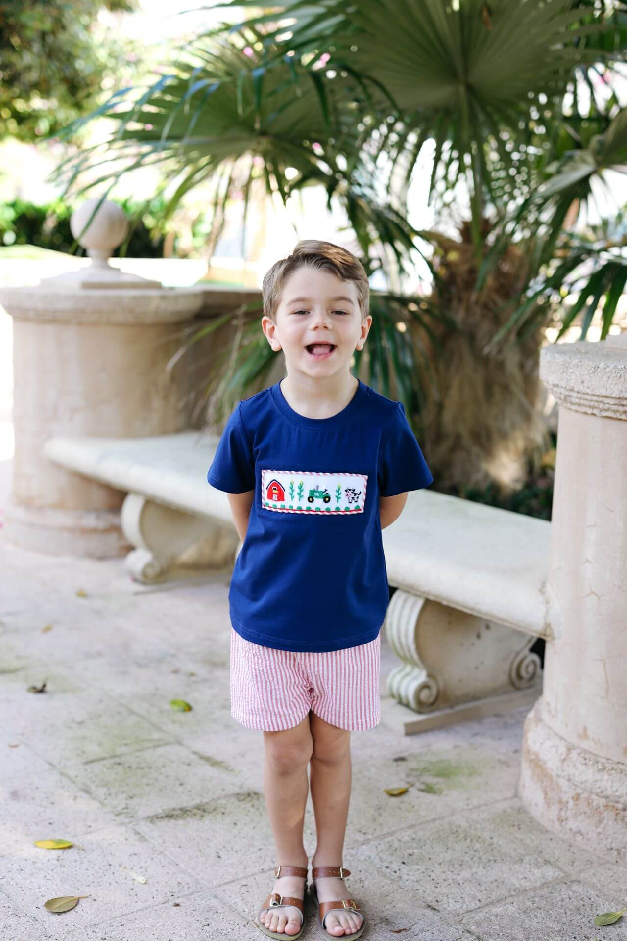 Child wearing a blue shirt with a graphic design, standing outdoors with greenery in the background