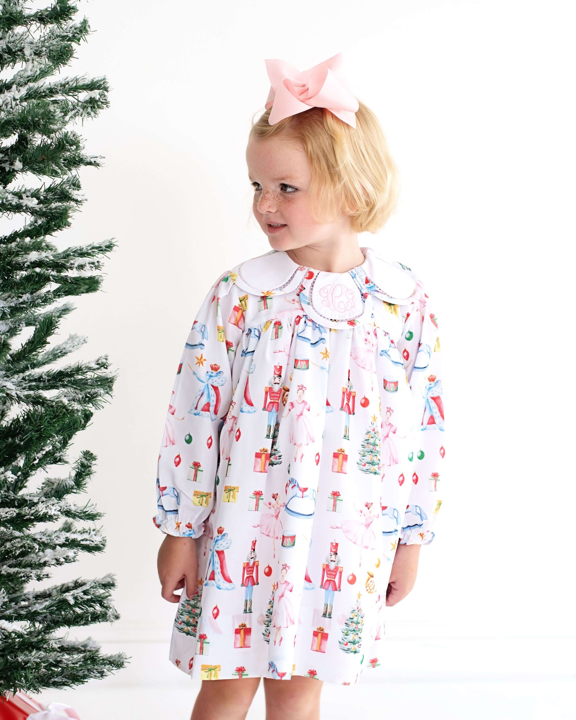 Child wearing a festive holiday dress with Christmas tree and presents pattern next to a decorated Christmas tree.
