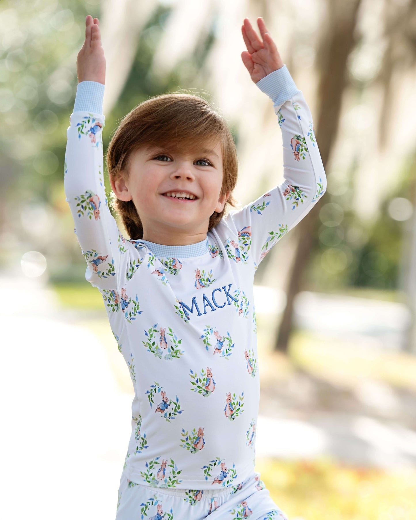 Child wearing a floral-patterned shirt with 'Mack' printed on it, standing outdoors with arms raised.