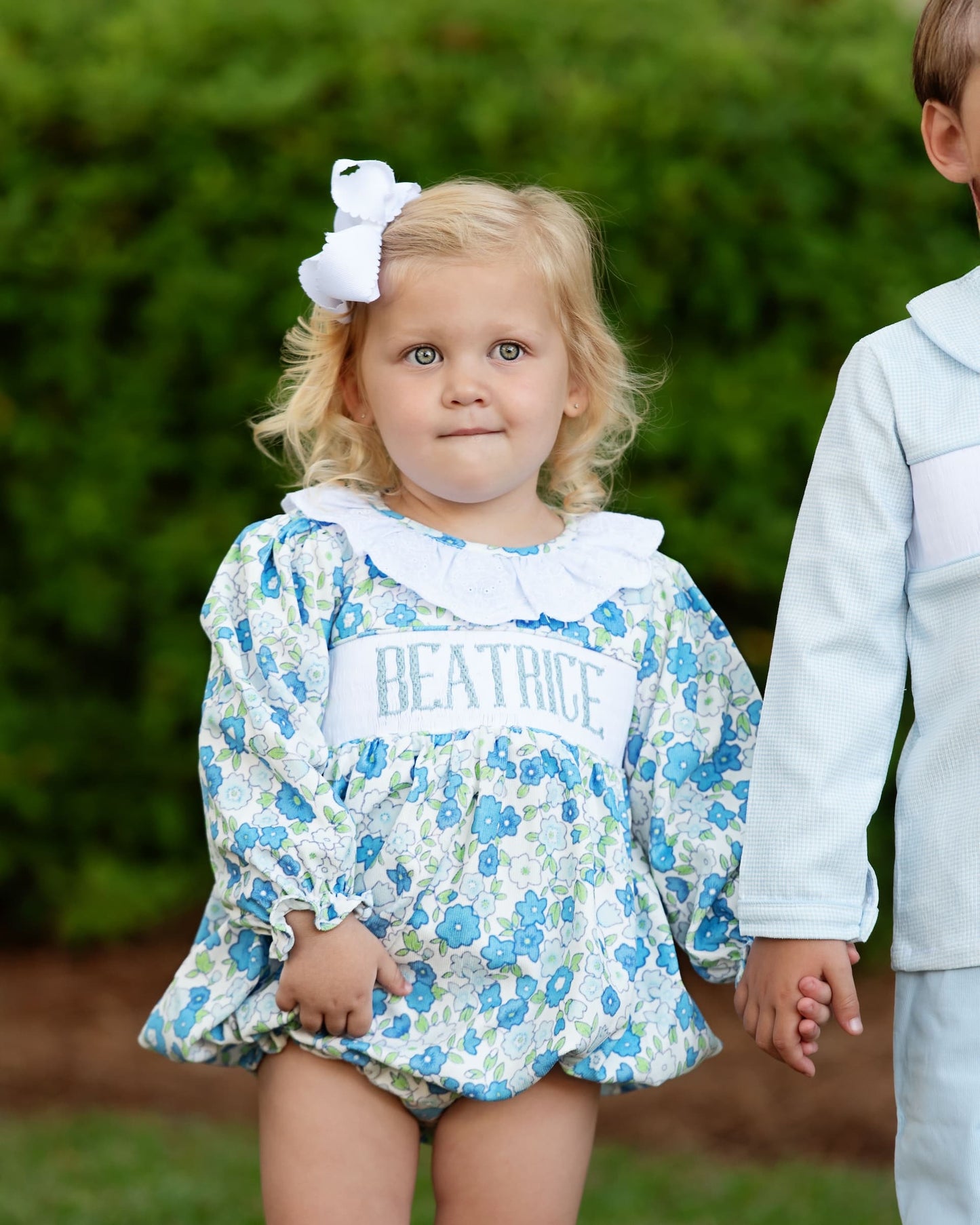 Child wearing a floral dress with 'BEATRICE' printed on it, standing outdoors.