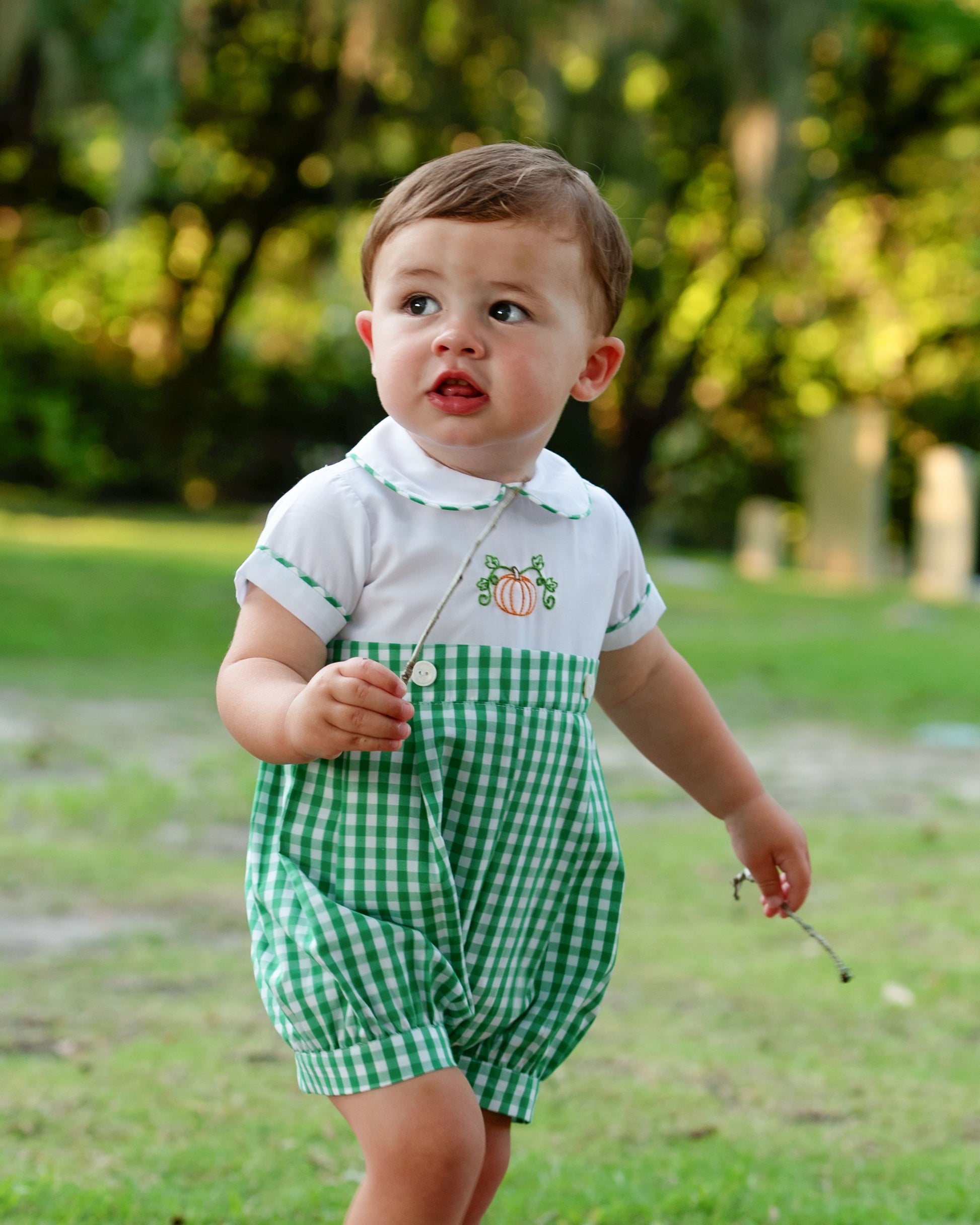 Young boy wearing a green checkered outfit with a white shirt featuring pumpkin designs, standing outdoors.