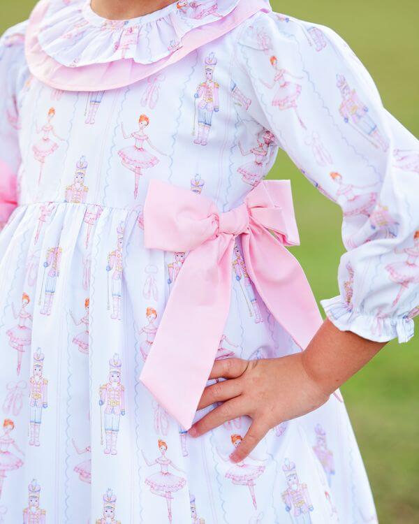 Child wearing a holiday print dress with a large pink bow, standing against a grassy background