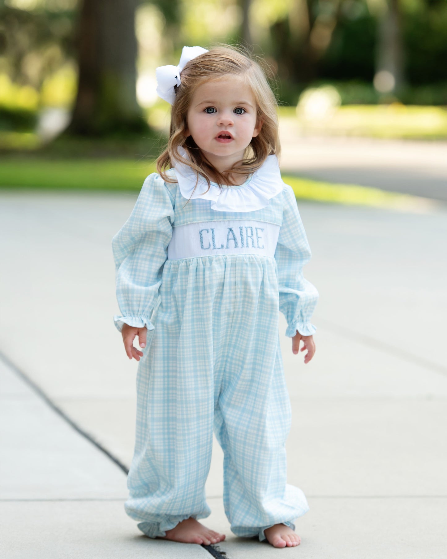 Child wearing a light blue checkered dress with 'CLAIRE' embroidered on it, standing outdoors.