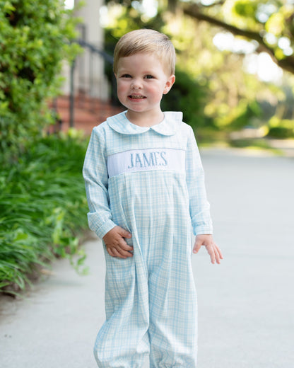 Child wearing a light blue checkered outfit with 'JAMES' printed on it, standing outdoors.