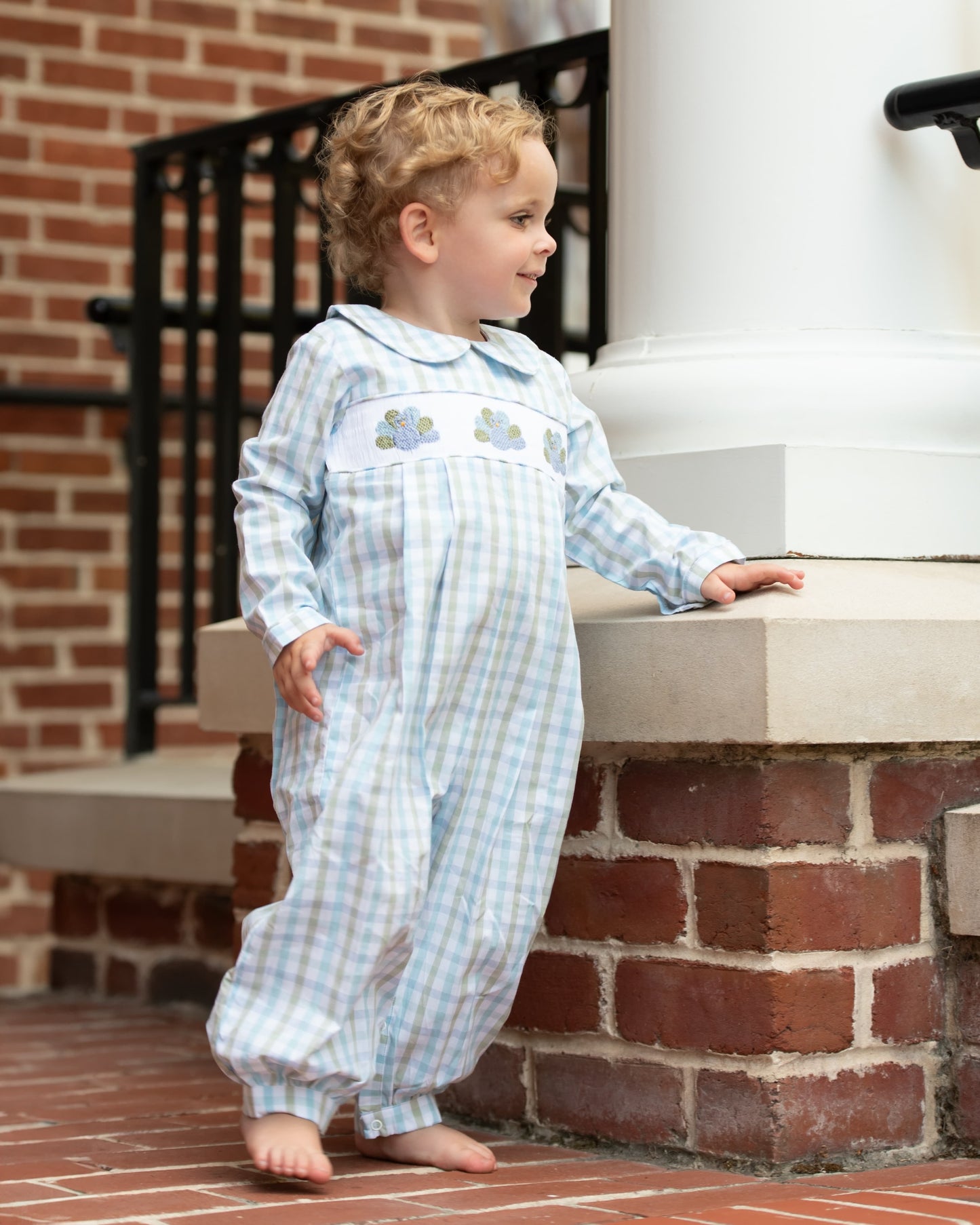 Child wearing a light blue checkered outfit with smocked turkeys standing on a brick step.