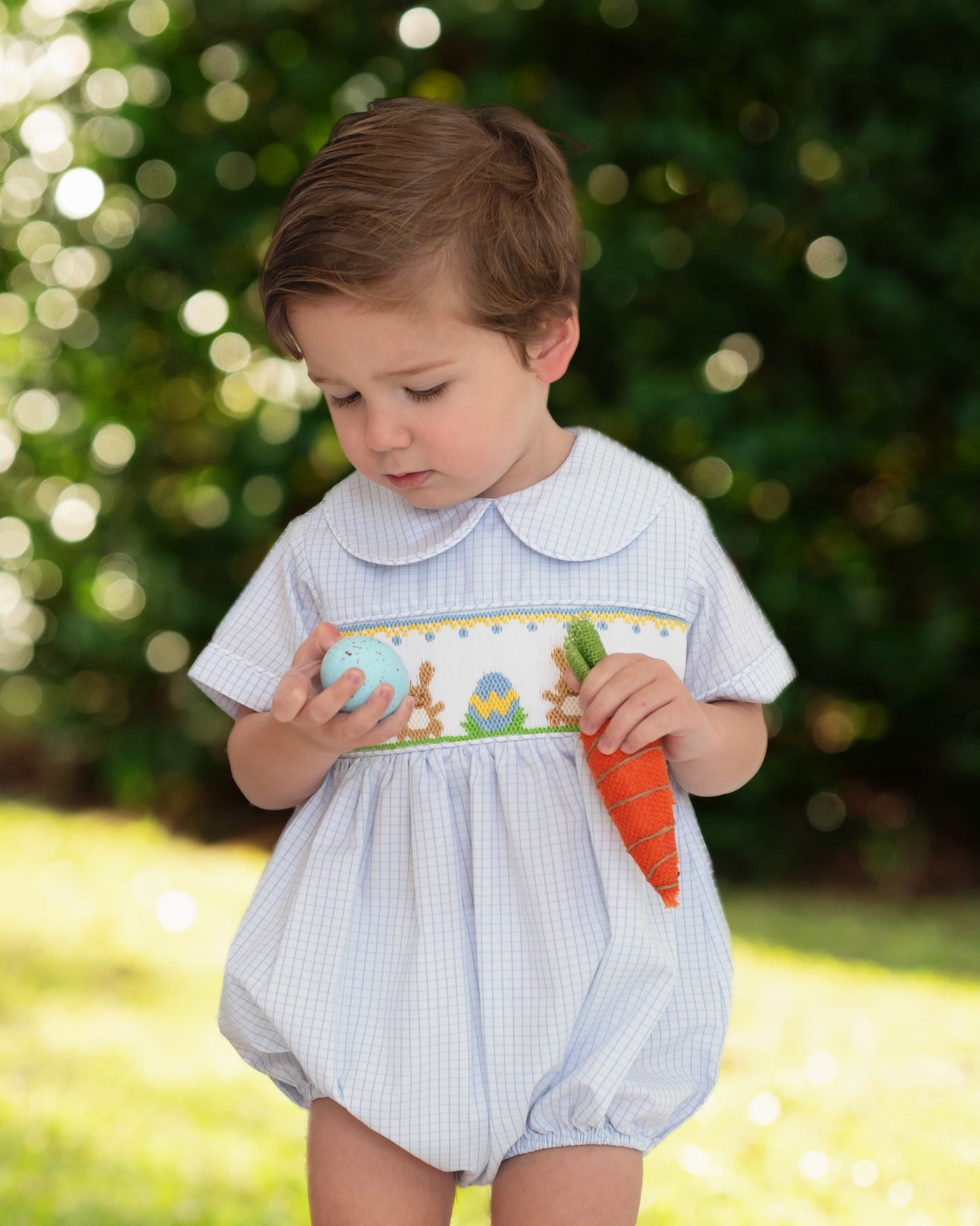 Child wearing a light blue checkered romper with a carrot, standing outdoors.