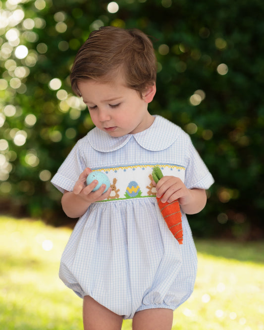 Child wearing a light blue checkered romper with a carrot, standing outdoors.