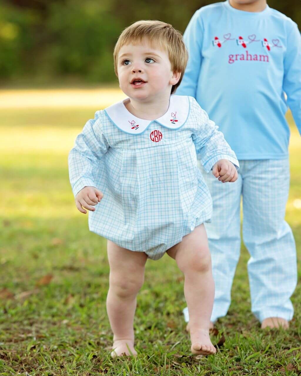 Child wearing a light blue checkered romper with a monogram, standing outdoors.