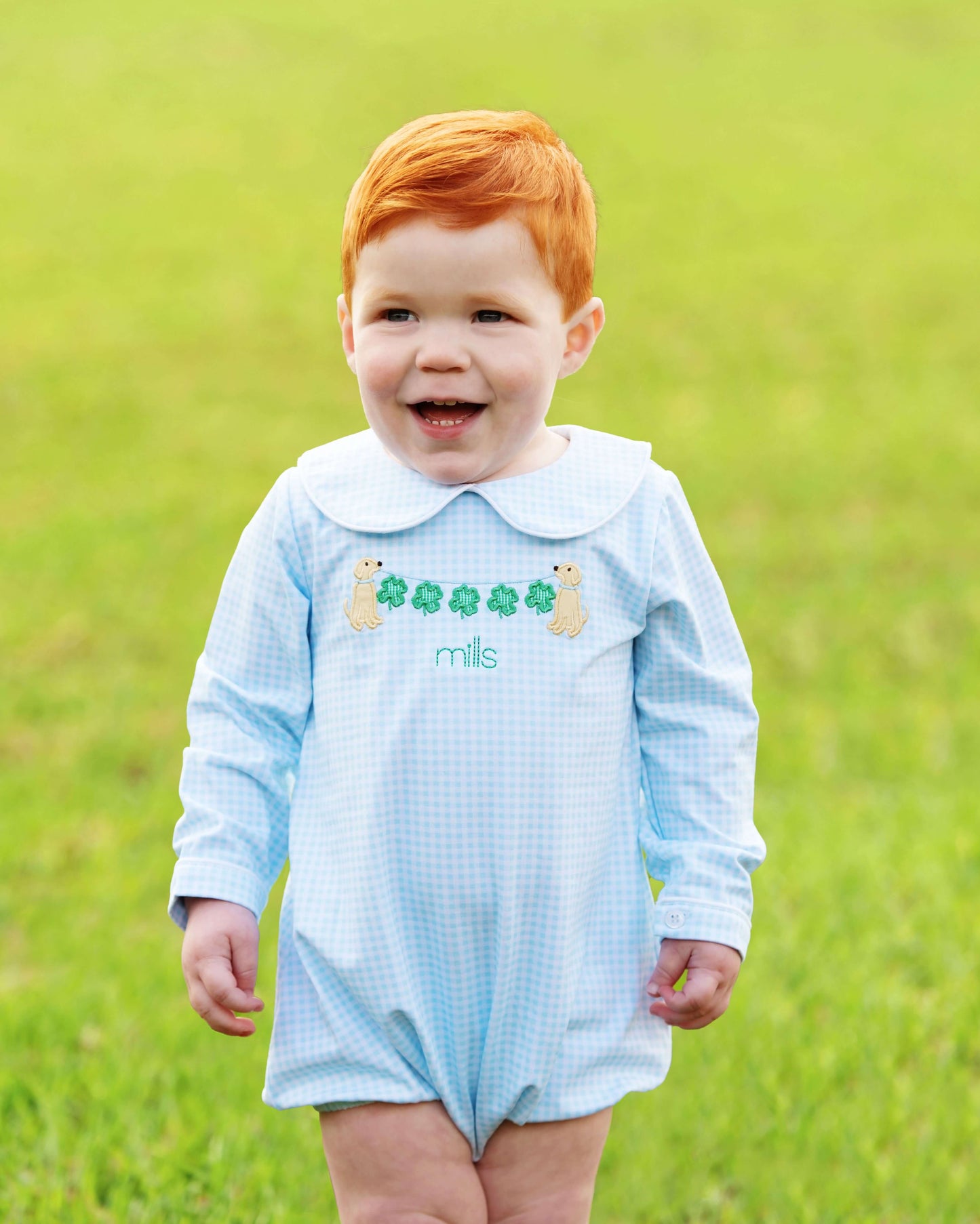 Child wearing a light blue checkered romper with embroidered design on a grassy background