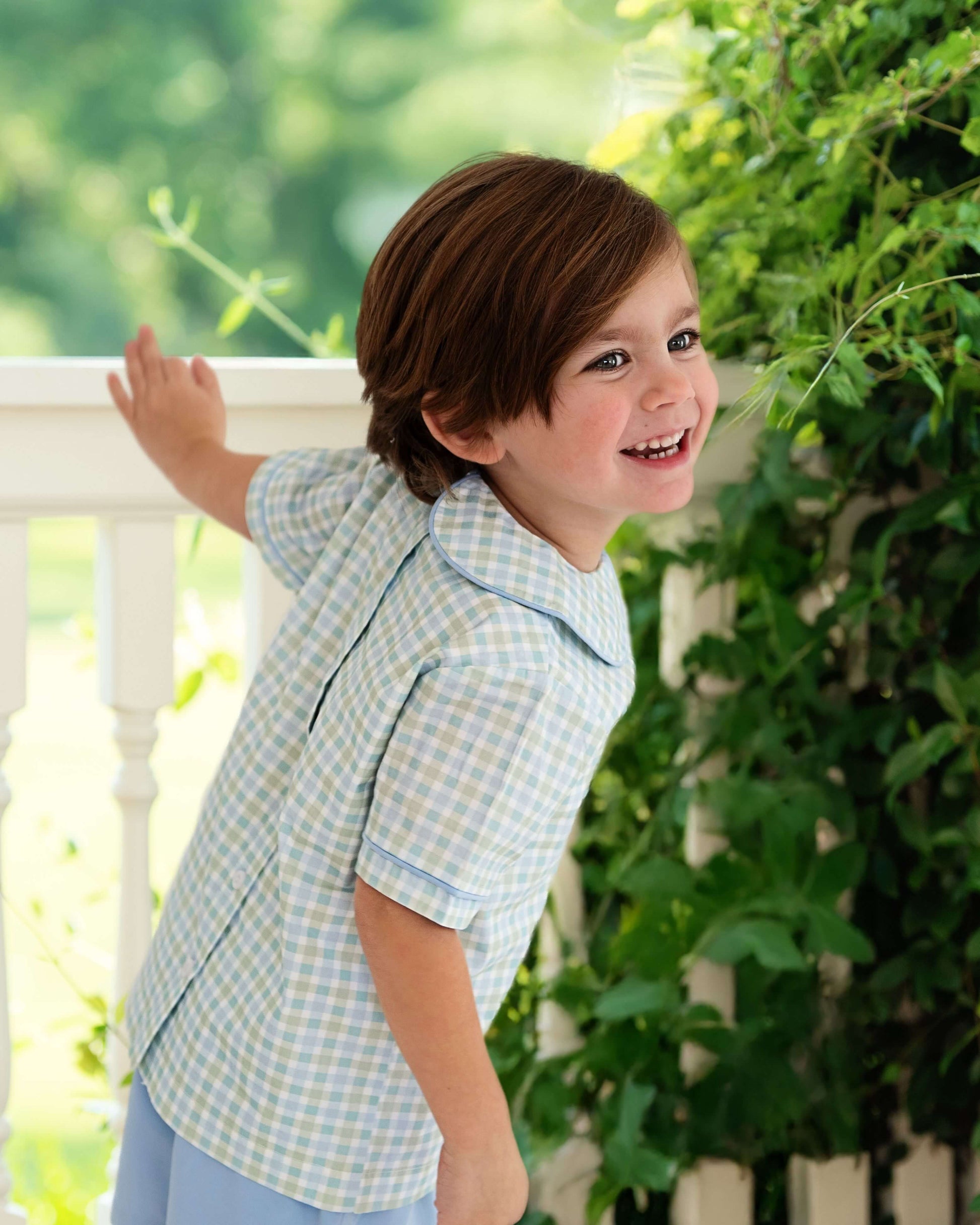 Child wearing a light blue checkered shirt standing outdoors with greenery in the background