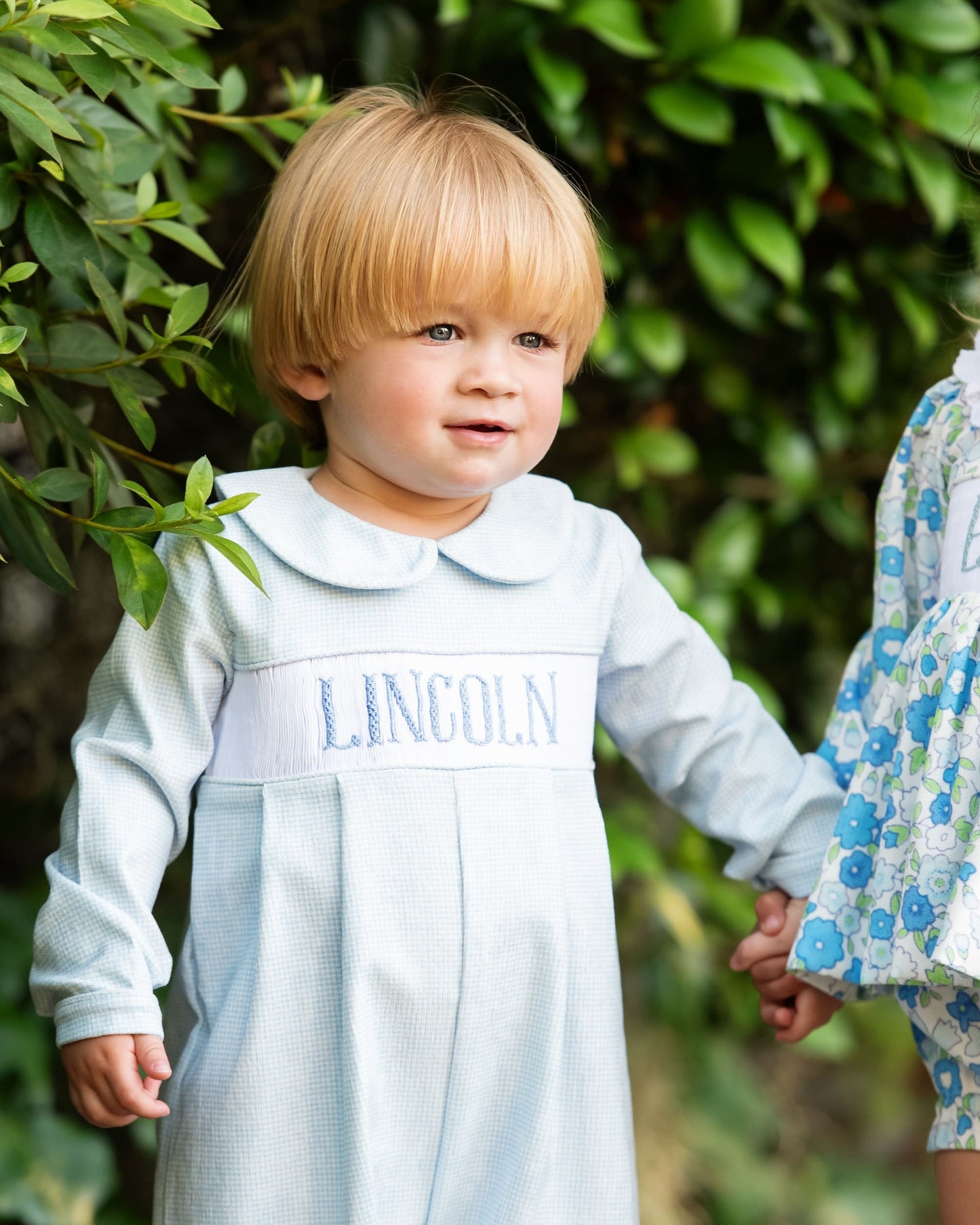 Child wearing a light blue dress with 'LINCOLN' embroidered on it, standing outdoors.