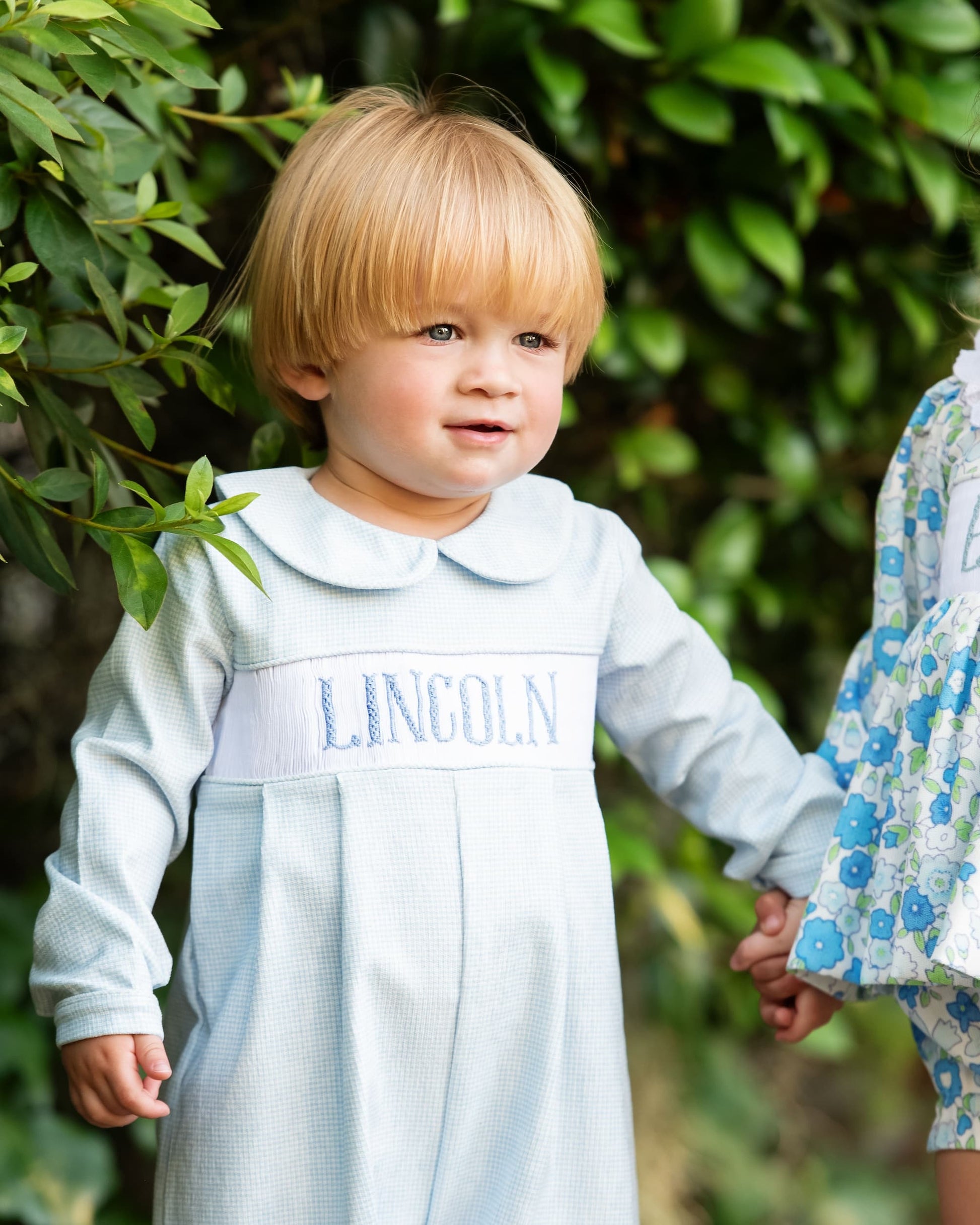 Child wearing a light blue dress with 'LINCOLN' embroidered on it, standing outdoors.