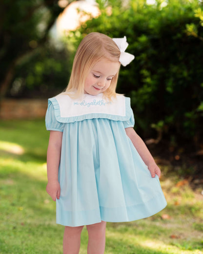 Child wearing a light blue dress with white trim and a white bow on her hair outdoors