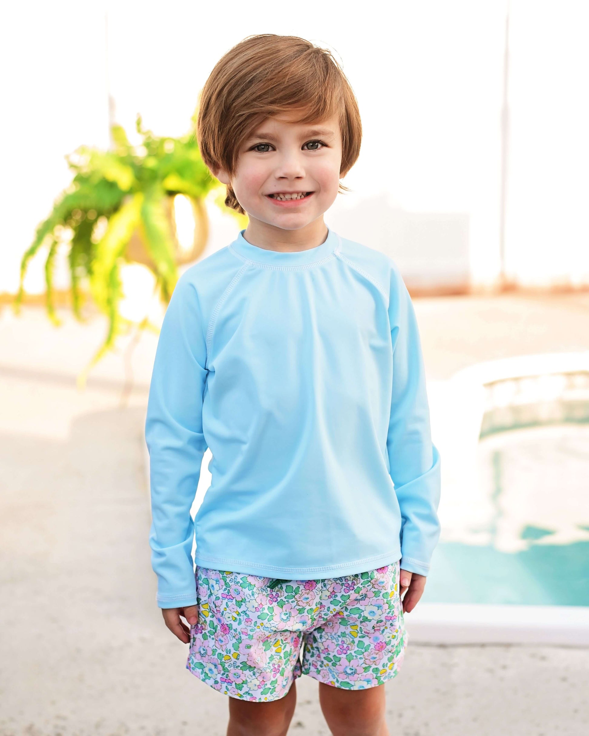 Child wearing a light blue long-sleeve shirt and colorful shorts by a poolside.