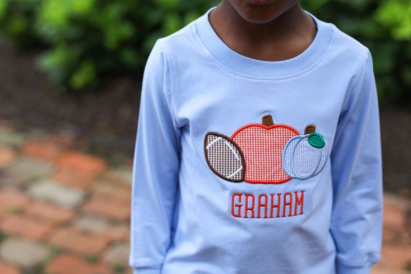 Child wearing a light blue long-sleeve shirt with a pumpkin design and name 'Graham' outdoors.