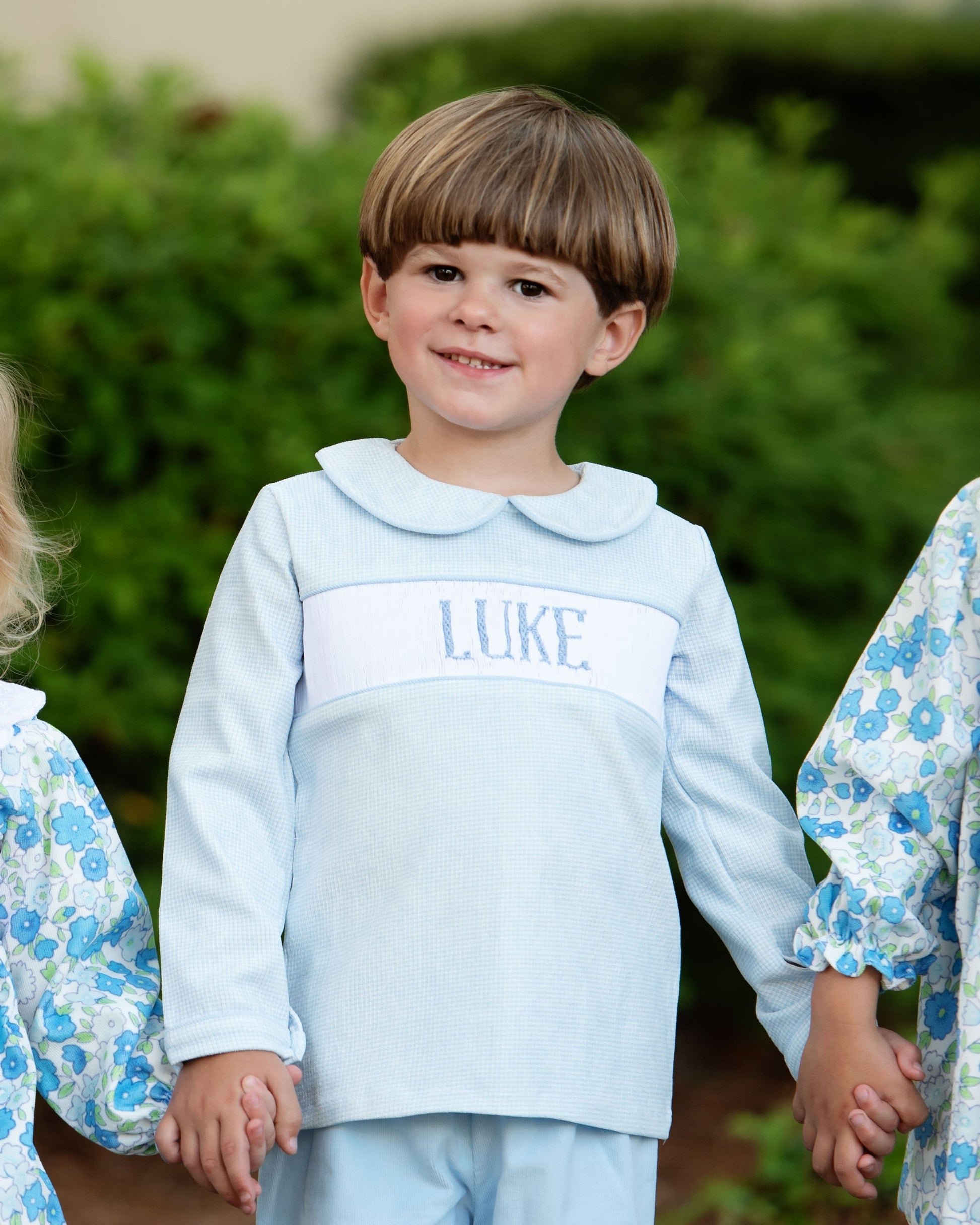 Child wearing a light blue outfit with 'LUKE' embroidered on it, standing outdoors with greenery in the background.