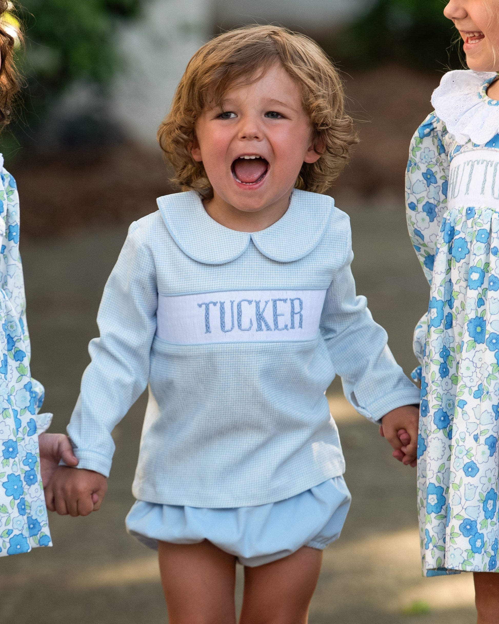 Child wearing a light blue outfit with 'TUCKER' on it, standing outdoors with other children.
