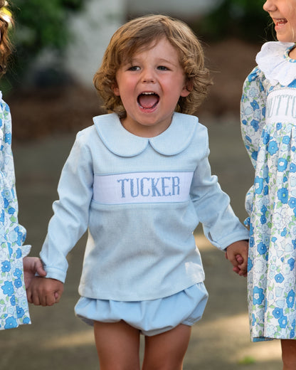 Child wearing a light blue outfit with 'TUCKER' on it, standing outdoors with other children.