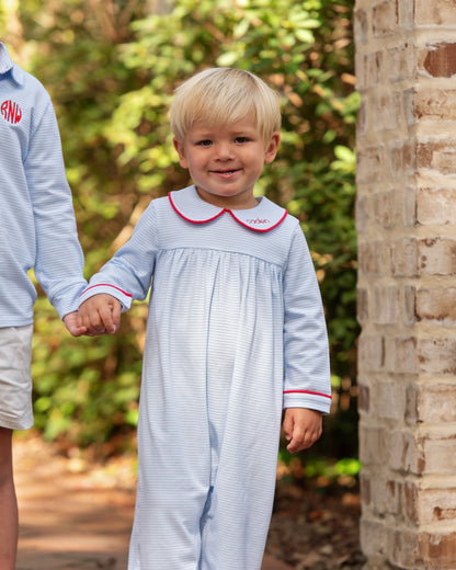 Child wearing a light blue outfit with a red collar, standing outdoors next to a stone wall.