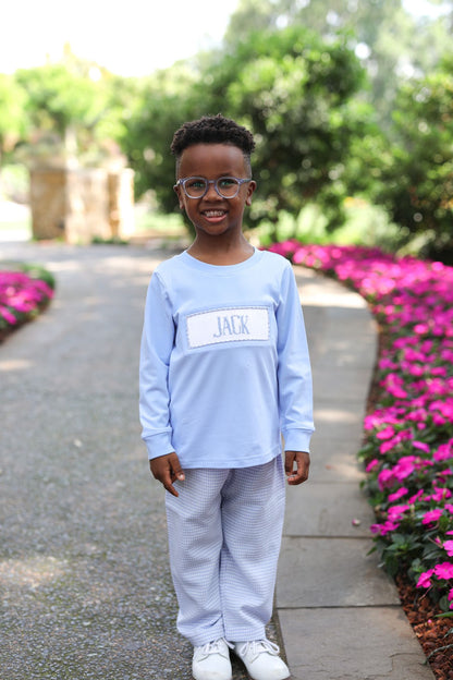 Child wearing a light blue shirt with 'JACK' printed on it, standing outdoors with pink flowers and greenery.