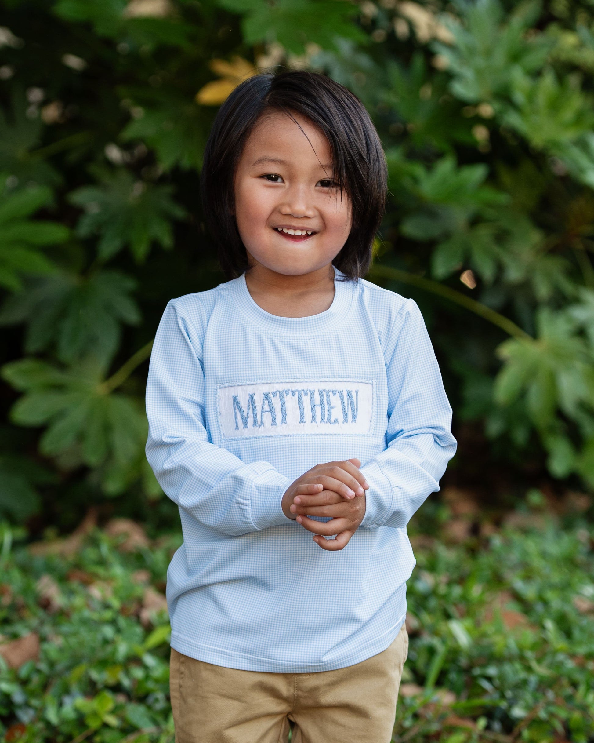 Child wearing a light blue shirt with 'MATTHEW' printed on it, standing in front of green foliage.
