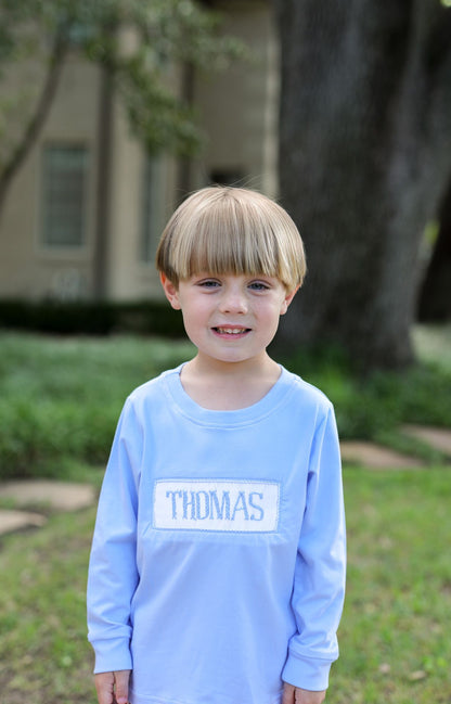 Child wearing a light blue shirt with 'THOMAS' printed on it, standing outdoors.