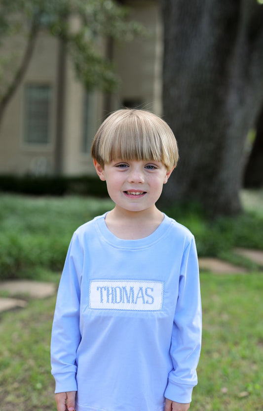 Child wearing a light blue shirt with 'THOMAS' printed on it, standing outdoors.