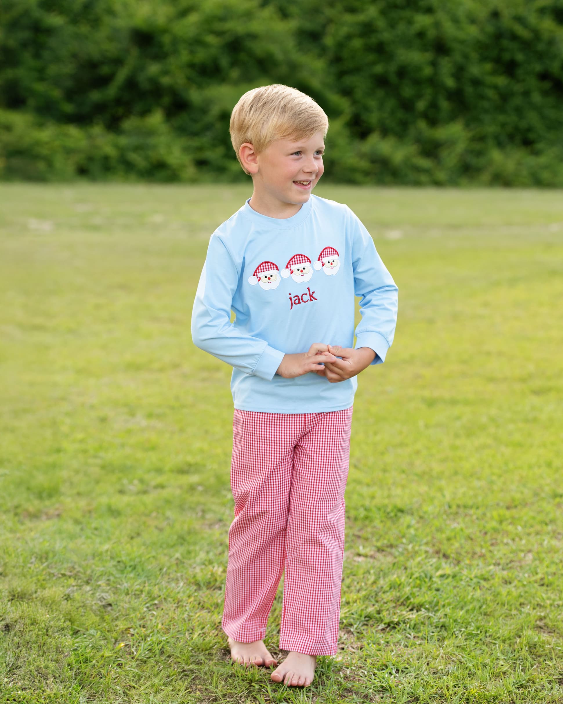Child wearing a light blue shirt with a snowman design and pink checkered pants standing in a grassy field.