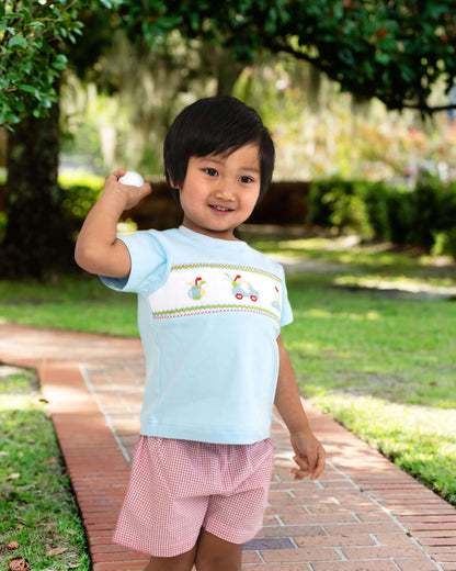 Child wearing a light blue shirt with embroidery and pink shorts standing on a brick path in a park.