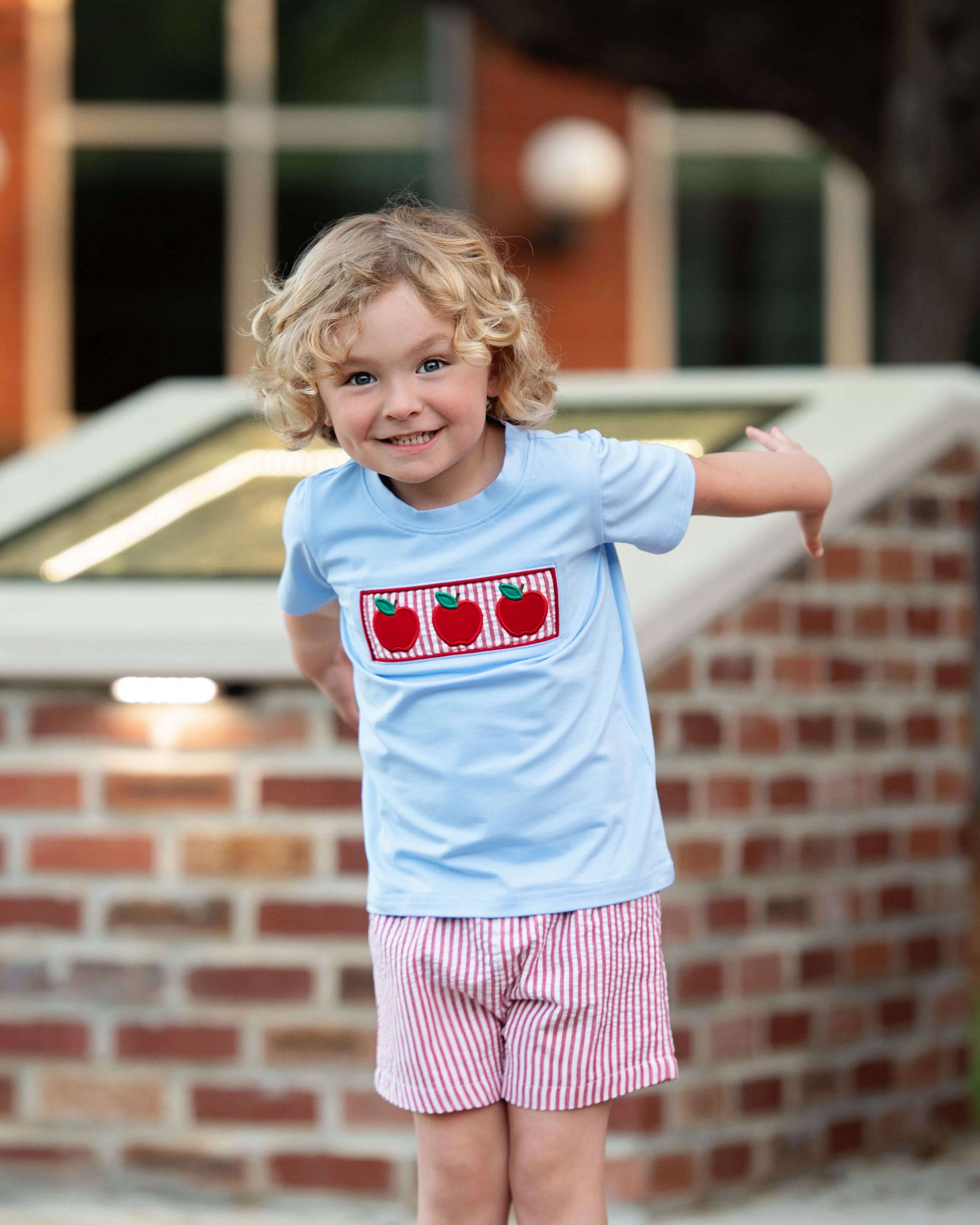 Child wearing a light blue shirt with red apple design and pink shorts standing in front of a brick wall.