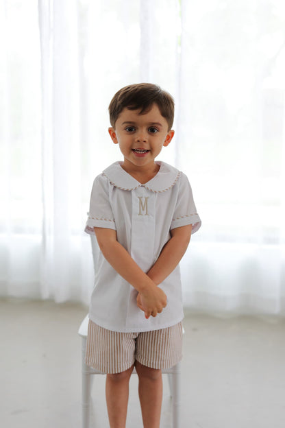 Child wearing a light gray shirt with a letter 'M' and beige shorts against a white curtain background