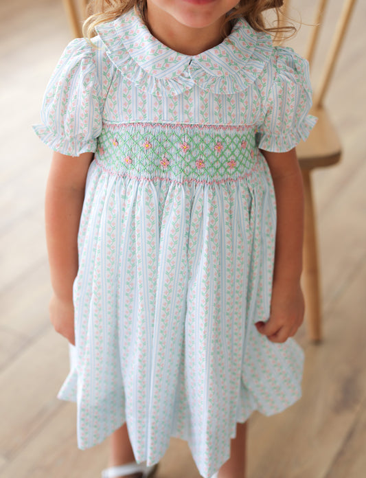 Child wearing a light green smocked dress with floral patterns on a wooden floor.