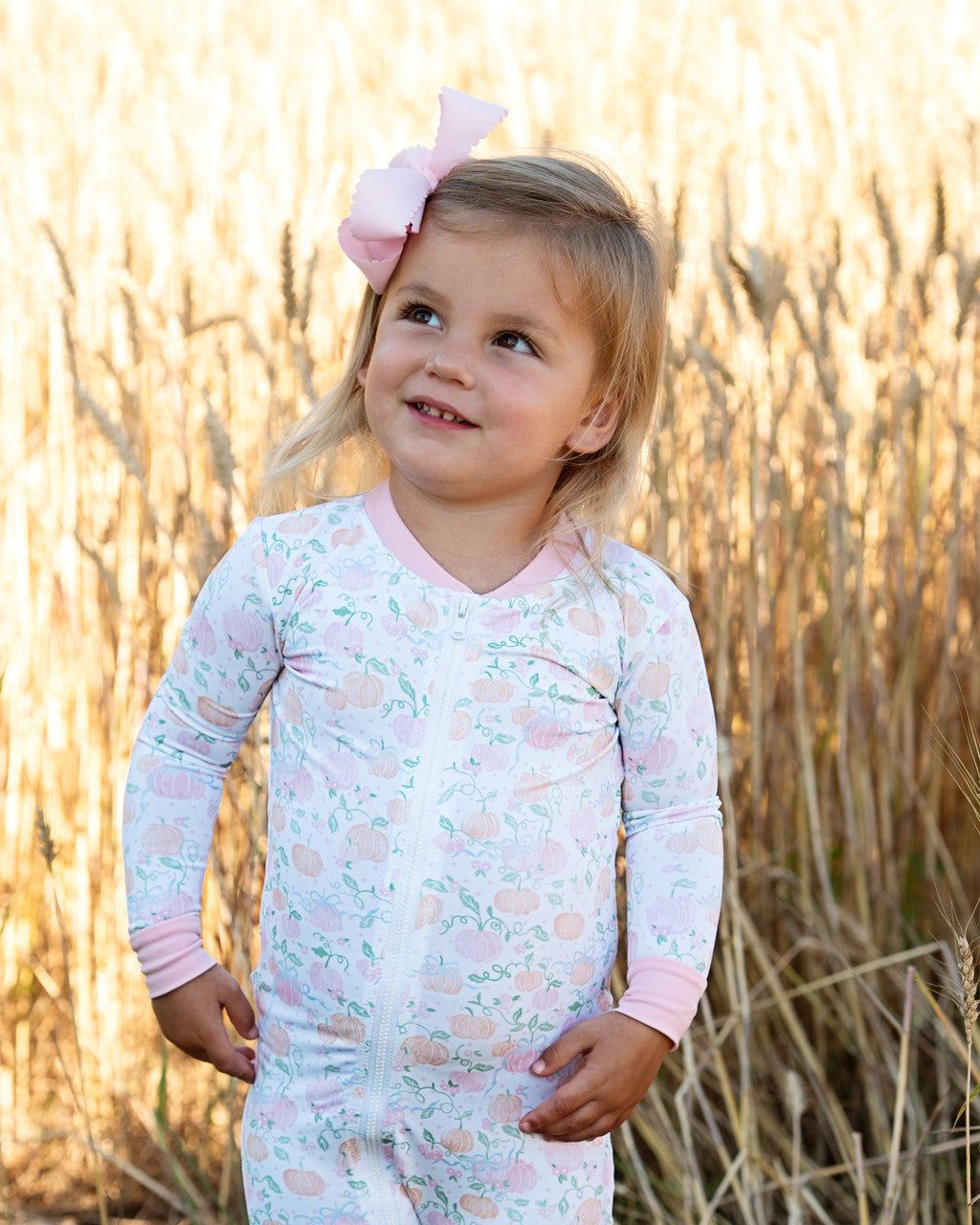 Child wearing a light pink and white pumpkin patterned outfit with a bow in a field.