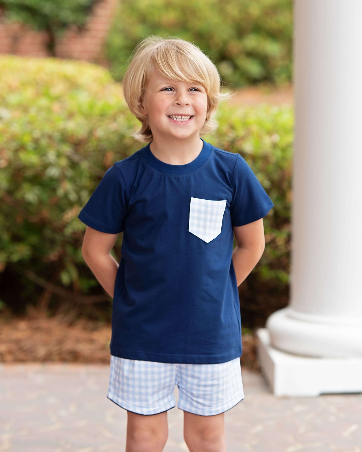 Child wearing a navy blue t-shirt with a white pocket and checkered shorts outdoors.