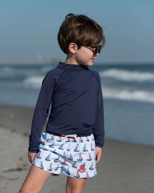 Child wearing a navy long-sleeve shirt and sailboat shorts on a beach.