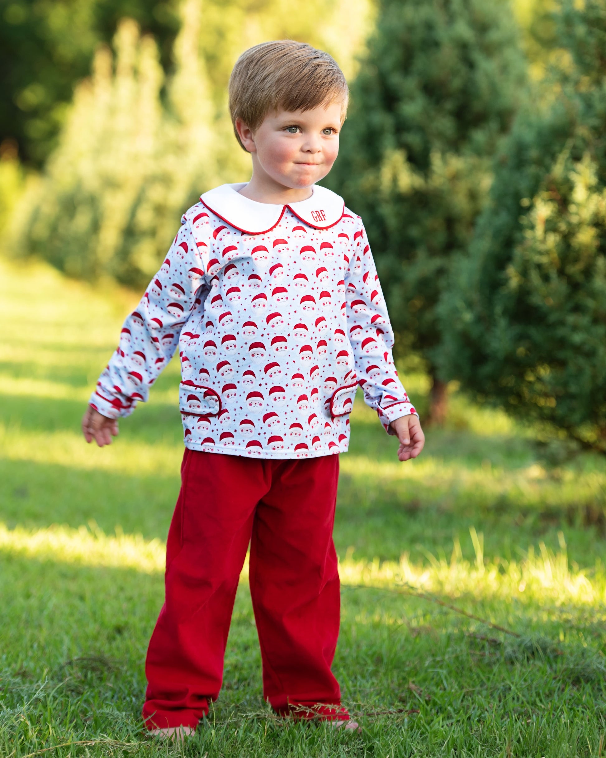 Child wearing a patterned shirt and red pants standing in a grassy area with trees in the background