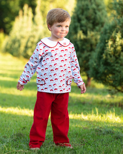 Child wearing a patterned shirt and red pants standing in a grassy area with trees in the background
