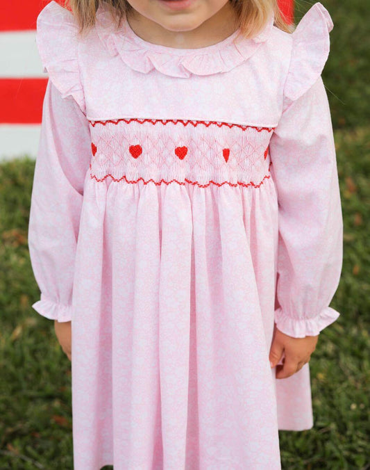Child wearing a pink dress with red heart details against a grassy background