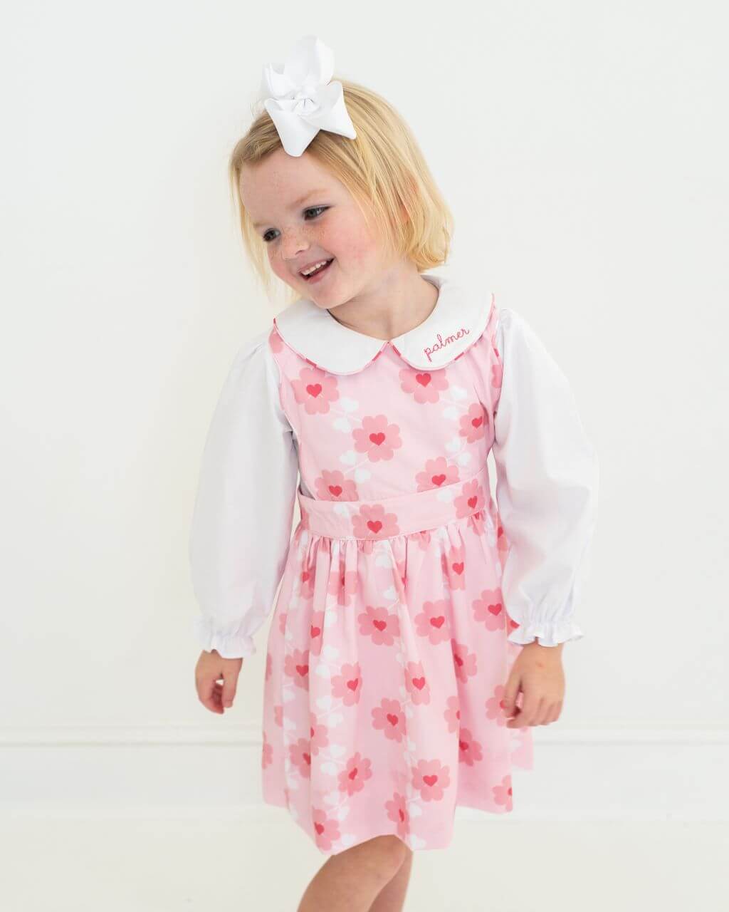 Child wearing a pink floral dress with a white collar and long-sleeve shirt on a white background