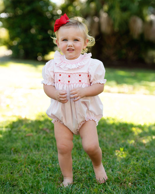 Child wearing a pink romper with ruffles and a red bow in an outdoor setting